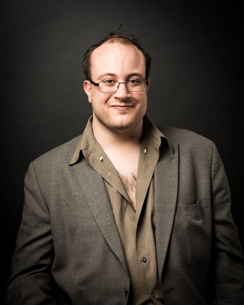 Alec Frazier, a tall, broad, brown haired, brown eyed man with a receding hairline and glasses smiles at the camera. He is wearing a business casual suit with an open collar.