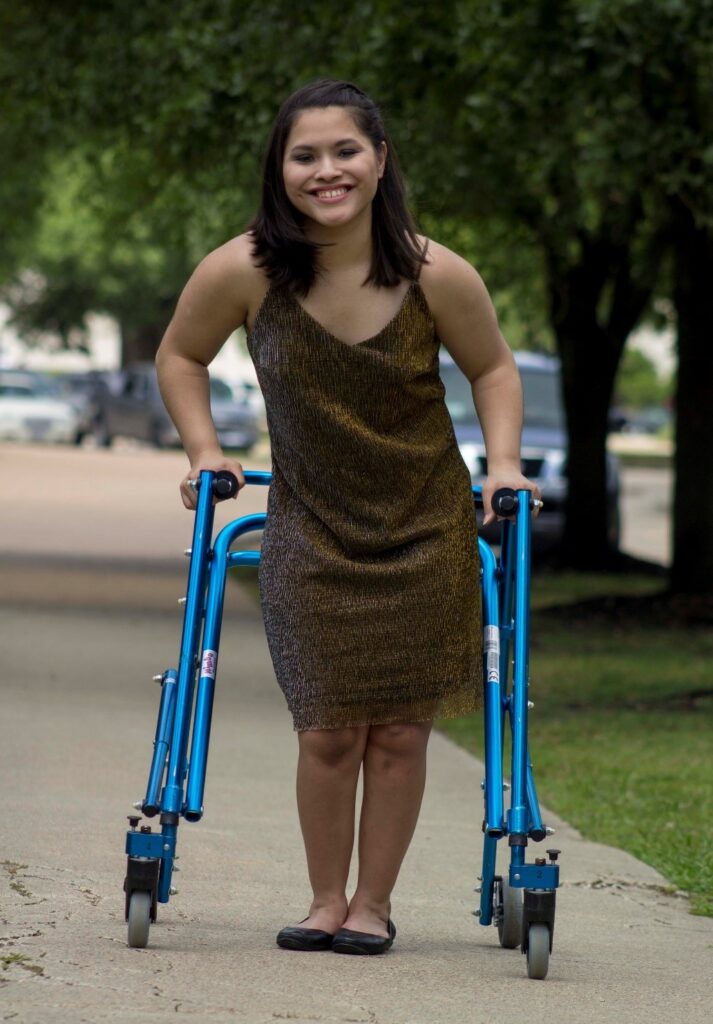 Alexa standing with her blue Walker wearing a gold strapless dress with black flats.