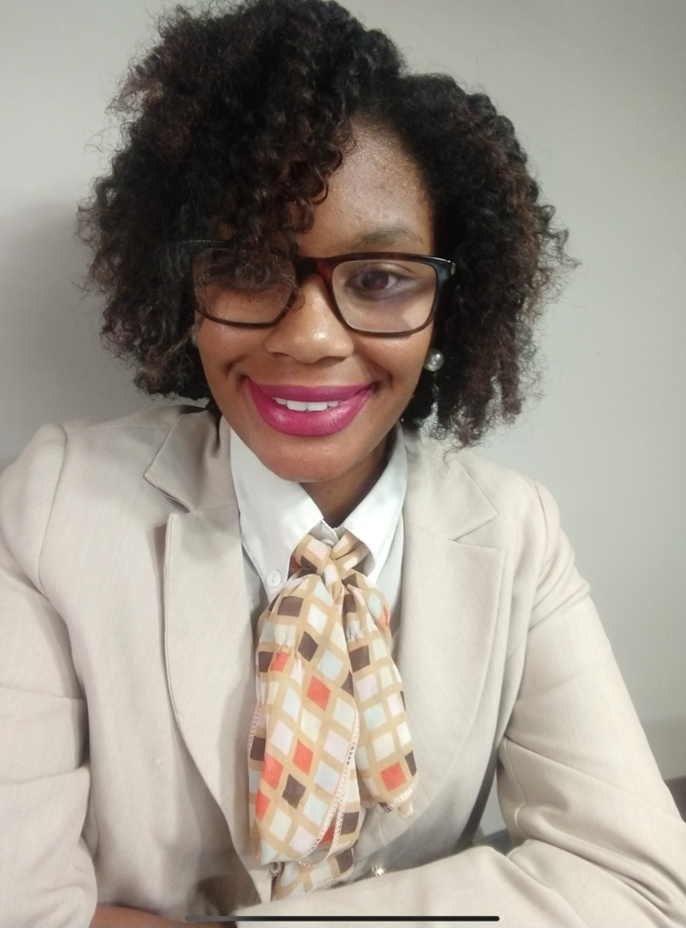 African American woman in front of a light background with glasses, pink lipstick and short curly hair, smiling at the camera with a light colored blazer on.
