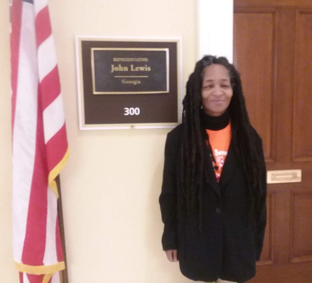 Anita Cameron, a disabled Black woman wearing an orange shirt, black turtleneck shirt, black suit jacket, and waist-length locs, stands smiling to the left of a dark wooden door. At the level of Anita’s right shoulder is a plaque on the wall that designates it as the door to the office of Representative John Lewis, of Georgia (room 300 in white letters underneath), with a U.S. flag, red and white stripes visible, to the right side of the sign.