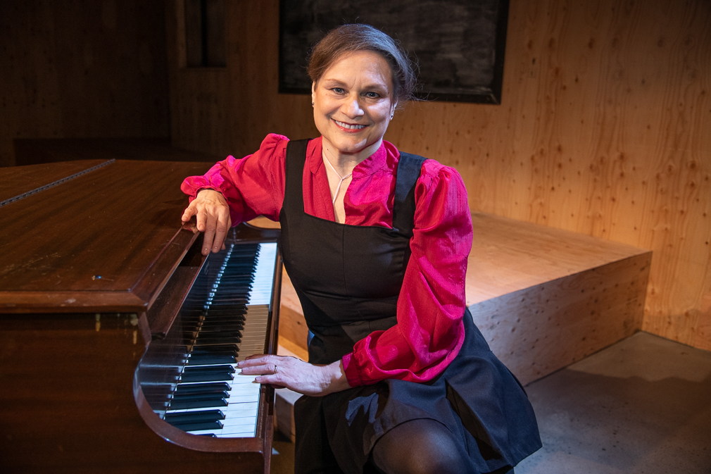 Caucasian face, blue eyes, short brown hair, sitting at piano with left had on keys and leaning on right elbow, wearing short black jumper with bright pink blouse.