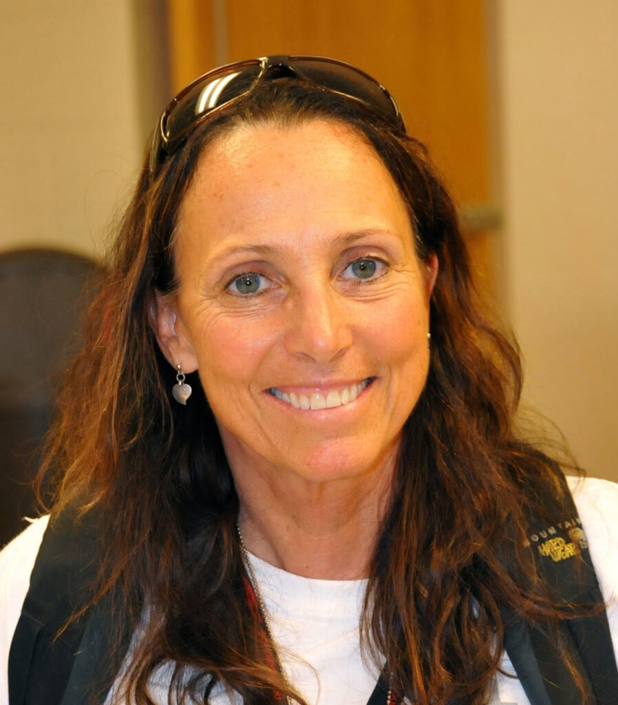 Caucasian woman with long brown hair, white shirt and vest, smiles at the camera.