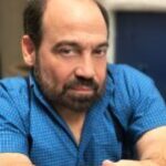 Caucasian man with a brown beard wears a blue shirt with a badass expression glares into the camera.