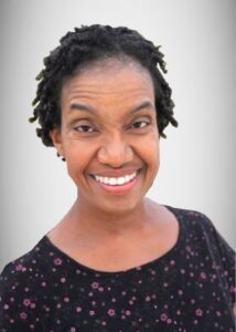 Head shot of African American woman in her 50's, facing forward and smiling, wearing a black shirt with small floral print, grey background.