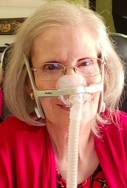 Photo of Diane Coleman wearing red print top and red sweater, smiling with gray bobbed hair, wire rimmed glasses and a nasal breathing mask.