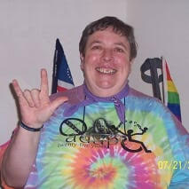 Fat, old, ruddy-white dyke with short brown hair,, big smile, tie-dyed ADAPT 25 rainbow tee shirt, sitting in power wheelchair, right hand displaying "I love you" sign.