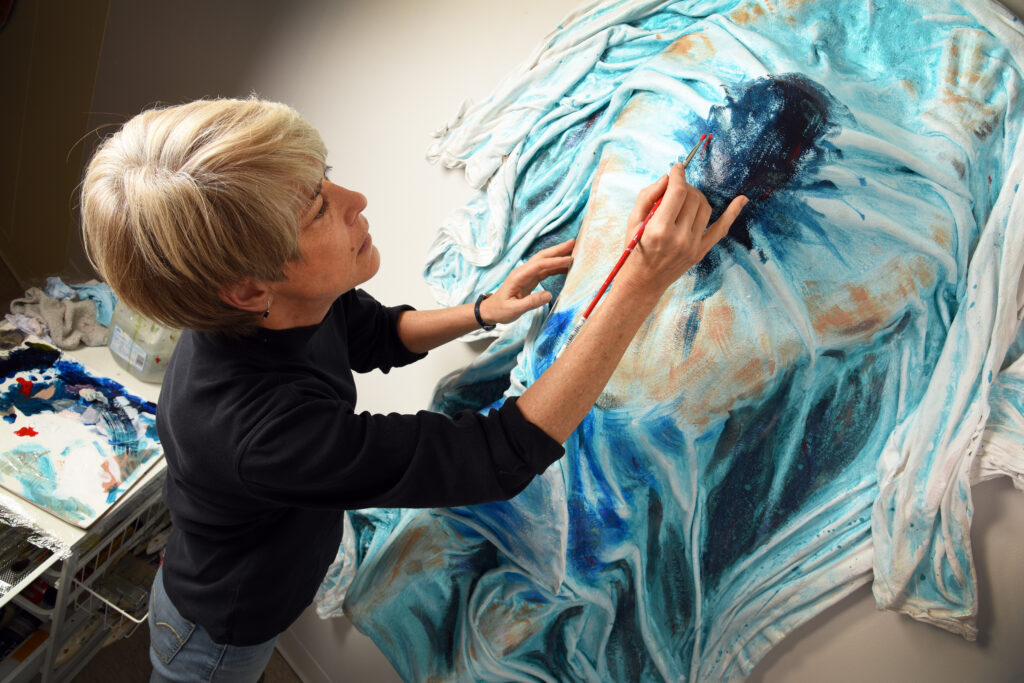 60 year old white female with short blondish hair and brown eyes, dressed in a black top and blue jeans. Seen from above painting a _gurative blue, white and skin tone sculpture of a small female paralympic swimmer.