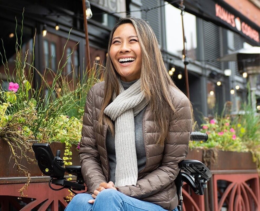 Happily smiling Asian girl with long hair sitting outside in a wheelchair at a sidewalk cafe with plants and flowers.