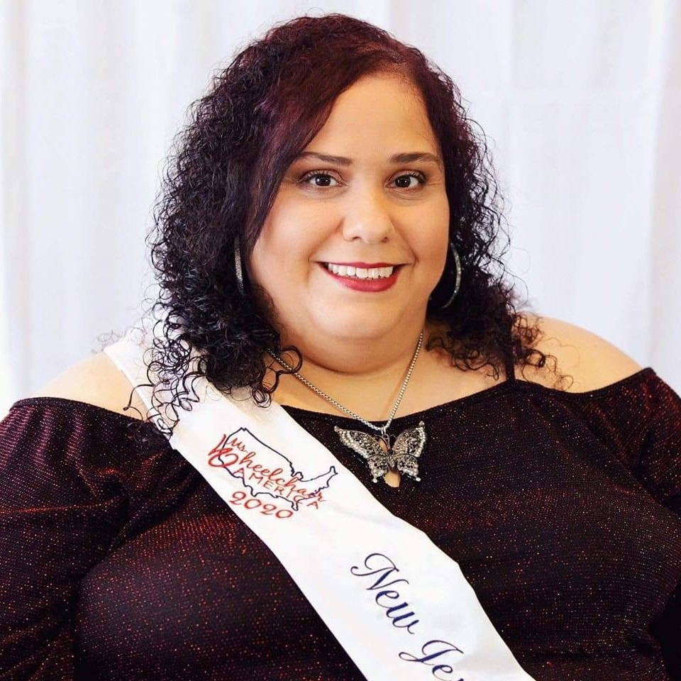 Millie Gonzalez, a plus-sized, Hispanic, disabled female with dark chocolate cherry-colored curly hair, wearing makeup, a large silver and black butterfly necklace, a dark red, glittery, cold-shoulder top and a sash that says Ms. Wheelchair America 2020-New Jersey, smiles at the camera..