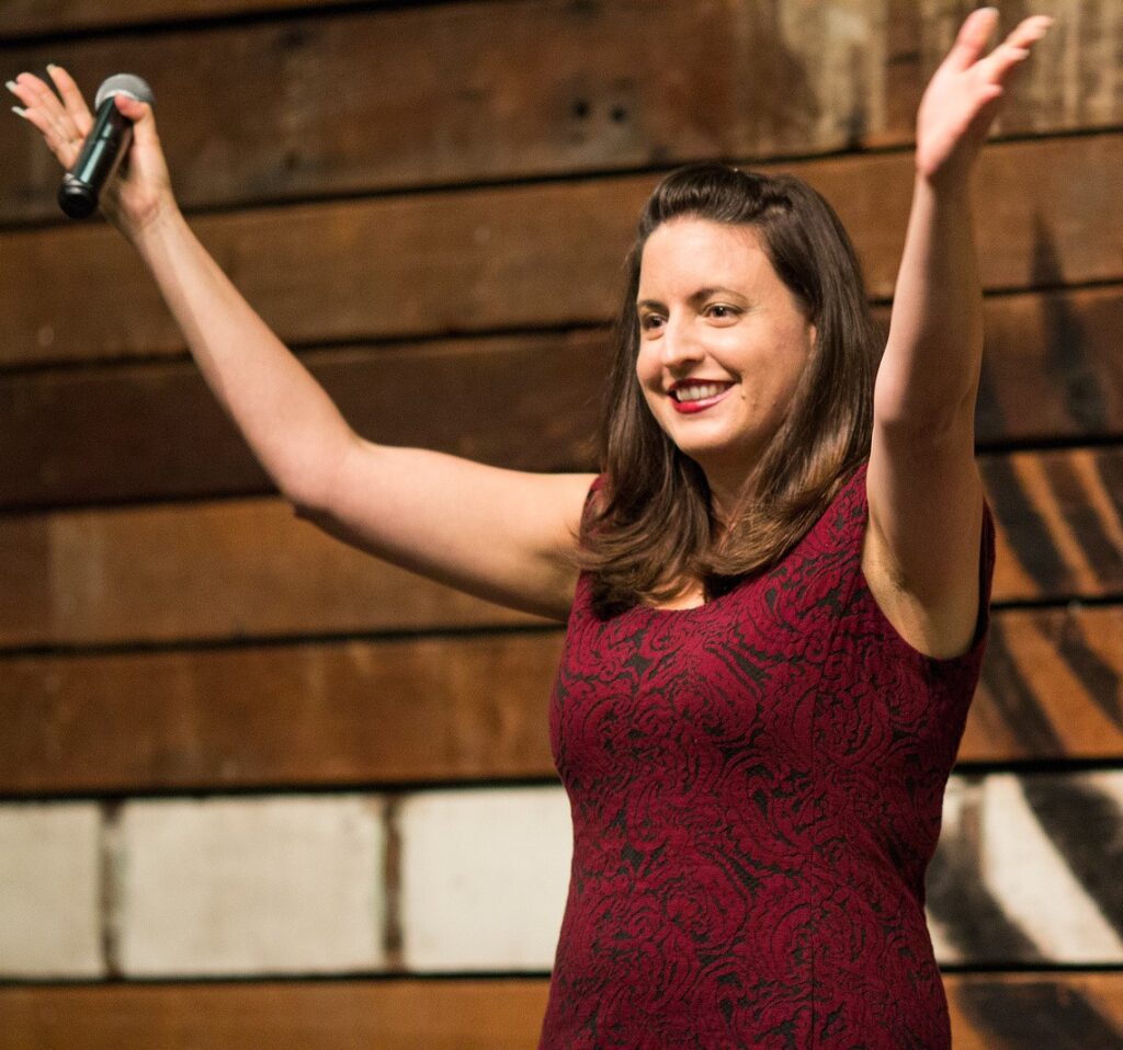 A white woman with brown eyes and long brown hair) raises her hands triumphantly with a microphone in one hand after telling a hilarious joke to an audience.