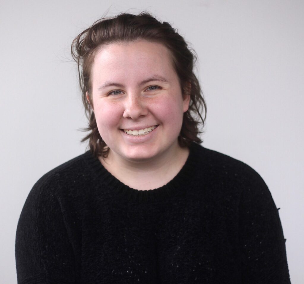 Headshot: Oaklee Thiele, a white female with short brown hair, wears a black sweater as she smiles looking directly at the camera.