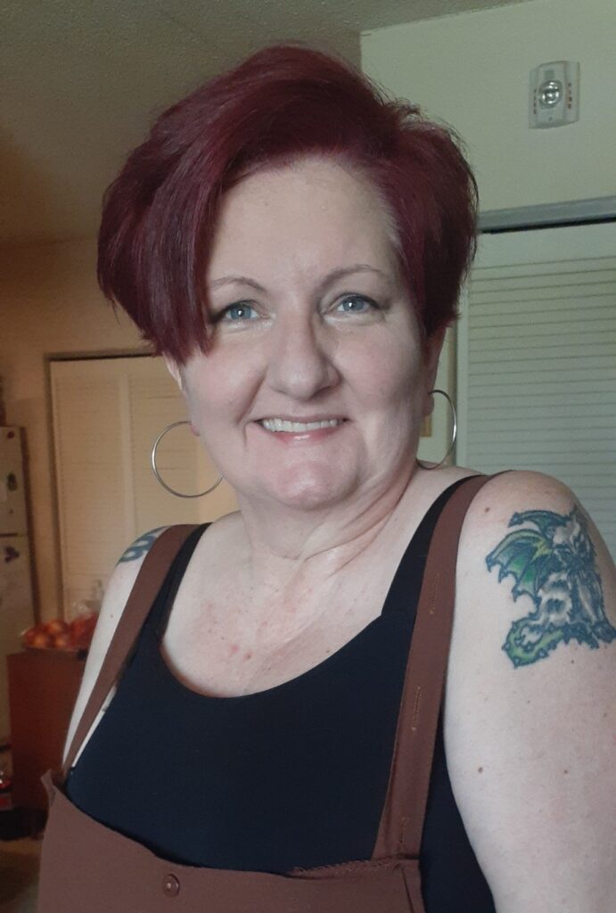 White, disabled woman standing with her hands in her pockets; with dark plum short hair, blue eyes, wearing a black tank top and brown overalls, and of course a smile.