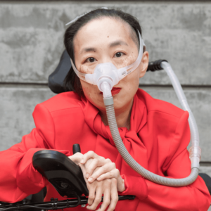Photo of an Asian American woman in a power chair. She is wearing an orange-red jacket and black pants. She is wearing a mask over her nose attached to a gray tube and bright red lip color. Her hands are resting over her joystick. Photo credit: Eddie Hernandez Photography