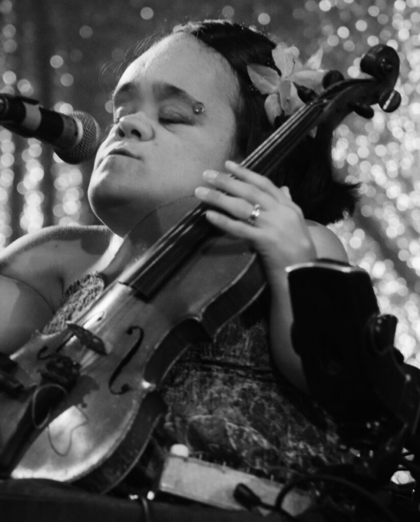 This is a black and white picture of Gaelynn Lea performing onstage from her electric wheelchair. She is a small, Caucasian woman with medium-length, dark hair. She has a flower clipped into her hair on the side of her head and she's wearing a patterned dress. The back wall of the stage is covered in sparkly sequins. There is a microphone in front of her. She is holding her violin upright like a tiny cello in her wheelchair. Her eyes are closed, and she deeply absorbed in the performance.