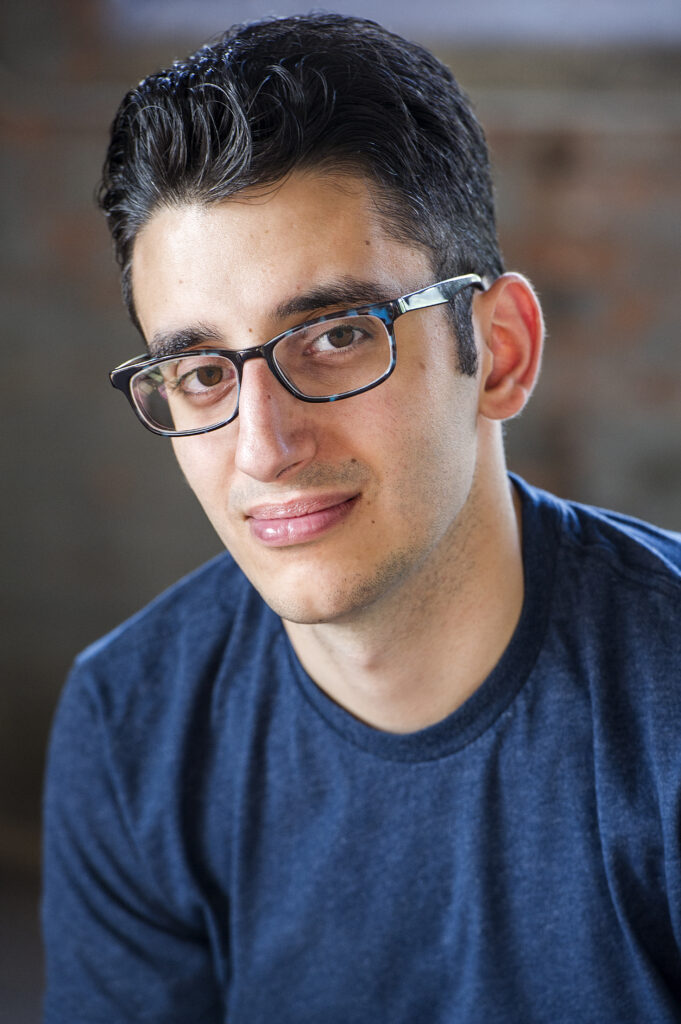 Photo of Ryan from the chest up. He is a man of Lebanese descent with brown eyes and short black hair that is slicked back. He wears black and blue tortoise shell glasses and a navy t-shirt. He gives a closed-mouth smirk and smiles with his eyes.