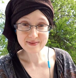 Smiling Jewish/multi-ethnic disabled woman, green leafy branches of trees in the daytime behind her. Her eyes are dark brown, her skin is light colored, and she has black rimmed oblong spectacles, pinched at the outer corners. She is wearing a black shirt underneath a looping paisley pattern in dark purples, and black earbuds with blue casing. Her head is wrapped with a fringed dark brown scarf.