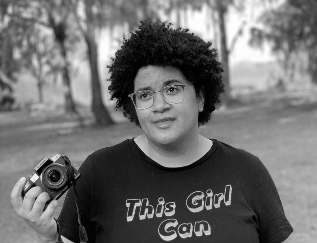 A bi-racial woman with gold glasses and short curly hair looking off to one side of the camera. She is holding a camera in her right hand. She is wearing a black shirt which says "This Girl Can"