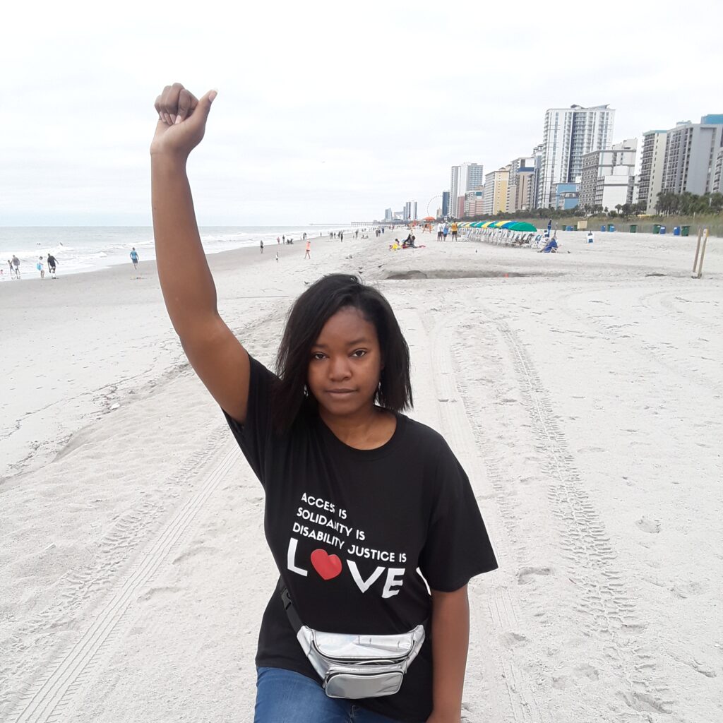 African American woman at the beach with her fist raised, wearing a black t-shirt with the words, “Access is Solidarity is Disability Justice is LOVE”