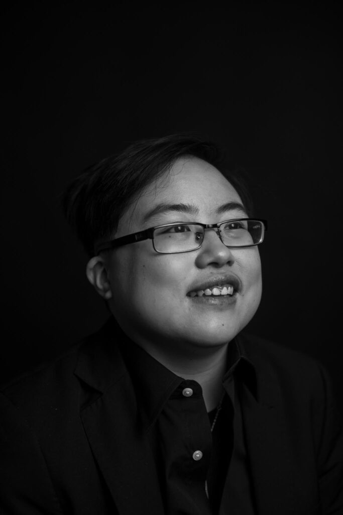 Black and white image of a young East Asian person with glasses smiling and laughing, looking slightly away from the camera. Photo by Colin Pieters.