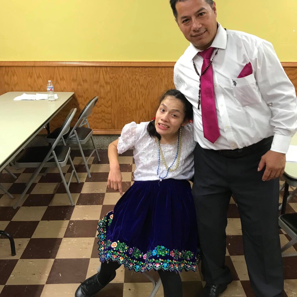 I’m with my uncle Julio, who is hugging me. We are smiling at the camera. I’m wearing my multicolored Ecuadorian clothes. He’s wearing a shirt with a pink tie and dress pants and has a rosary around his neck.