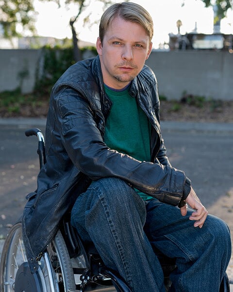 A blonde gentleman sits in a manual wheelchair and wears a leather jacket over a green shirt, outside during the day.