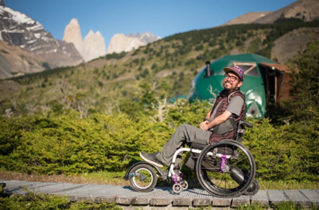 Caucasian male wheelchair user wearing brown sneakers, pants, shirt and vest smiles in front of an outdoor scene of green hills.