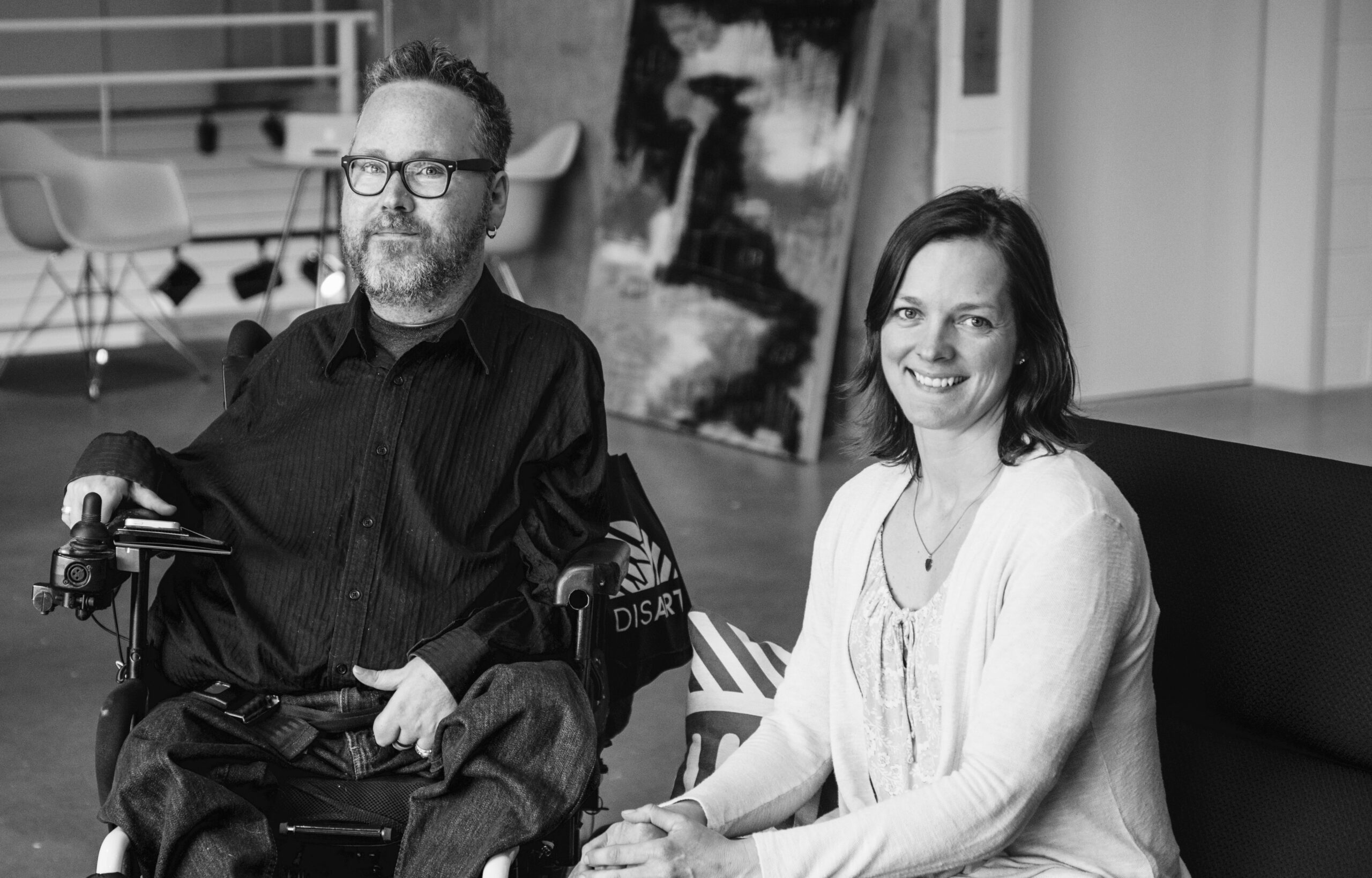 Black and white photo of Caucasian couple sitting next to each other smile at the camera. The man is sitting in his power wheelchair, holding the joy stick.