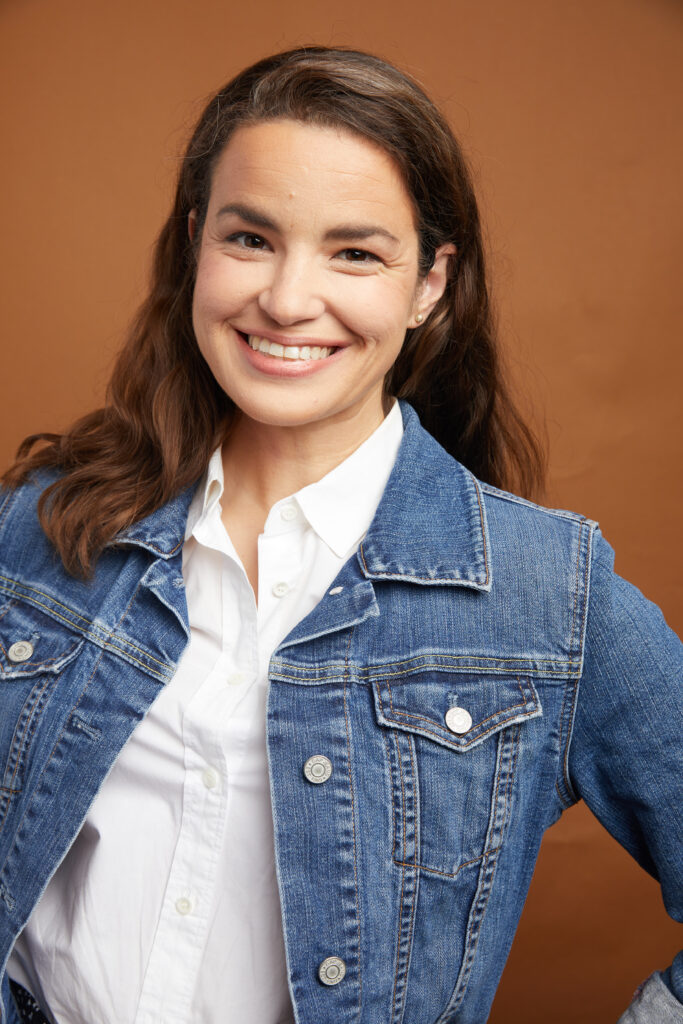 Light skinned woman with brown hair pushed past her shoulders wearing a white button down shirt and a denim jacket smiling at the camera.