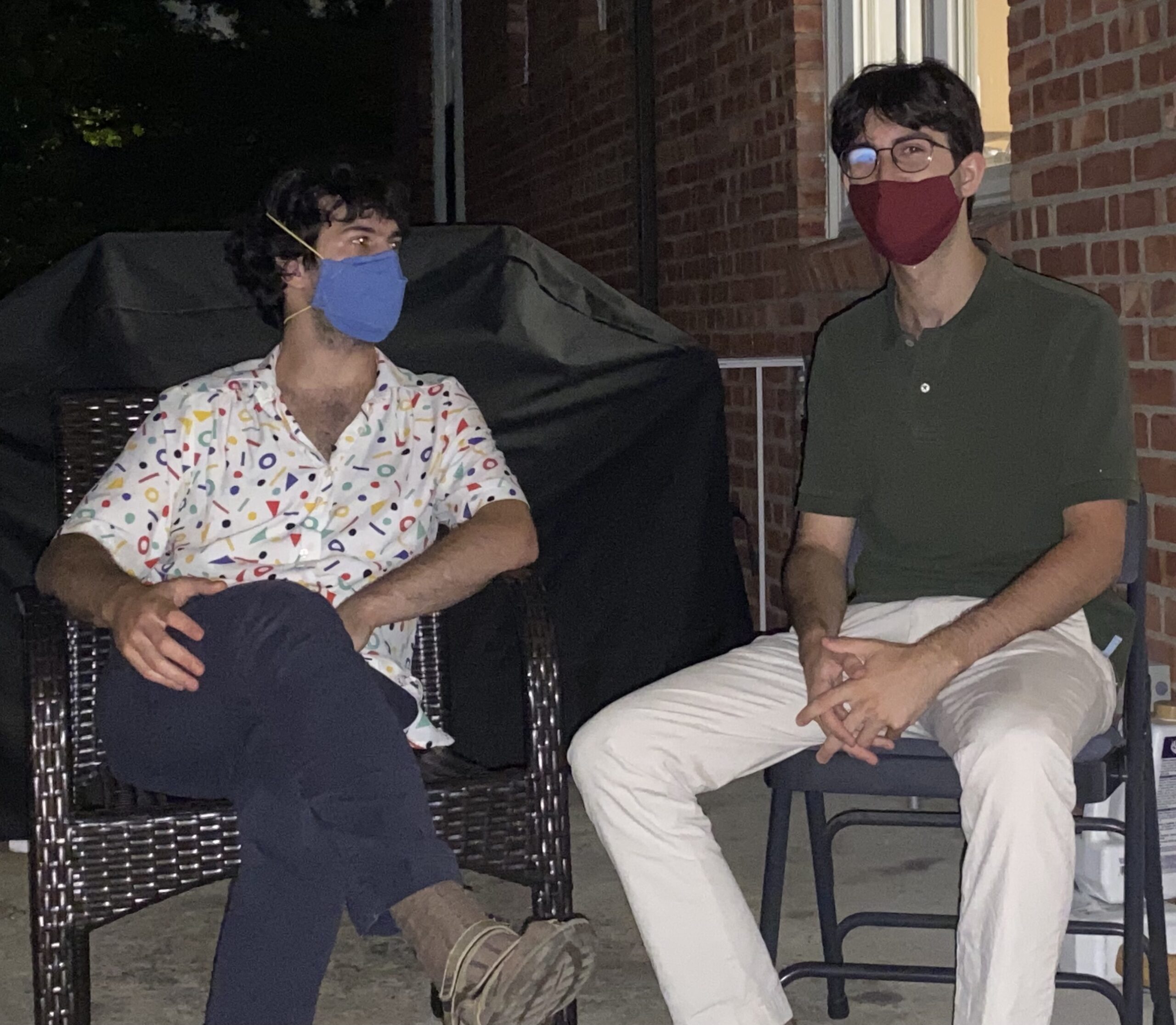 Two white Ashkenazi Jewish disabled brothers with dark brown hair sitting on a porch off of a red brick home on a summer night, dark sky in the background. Ezra is wearing a blue face mask, short sleeve white shirt with red, green, blue, yellow geometric shapes with blue pants, legs crossed and looking at Noah. Noah is wearing a red face mask, round metal glasses, a green short sleeve polo shirt, white pants with hands crossed in lap and legs spread, head looking straight towards the camera. Photo credit Shai Katz