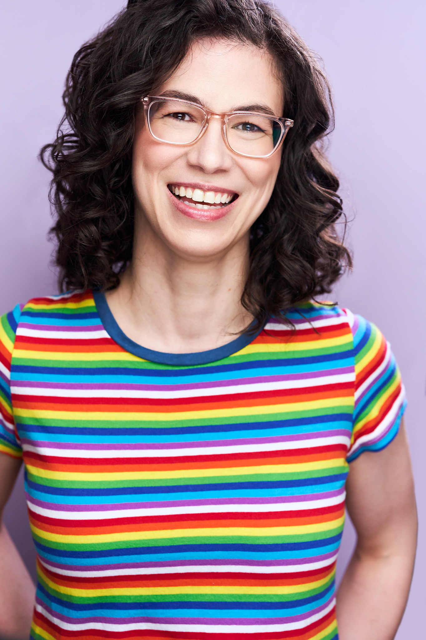 A female-presenting person with pale skin, clear plastic glasses, and shoulder-length curly brown hair, wears a rainbow striped T-shirt. They are smiling and standing against a light purple background