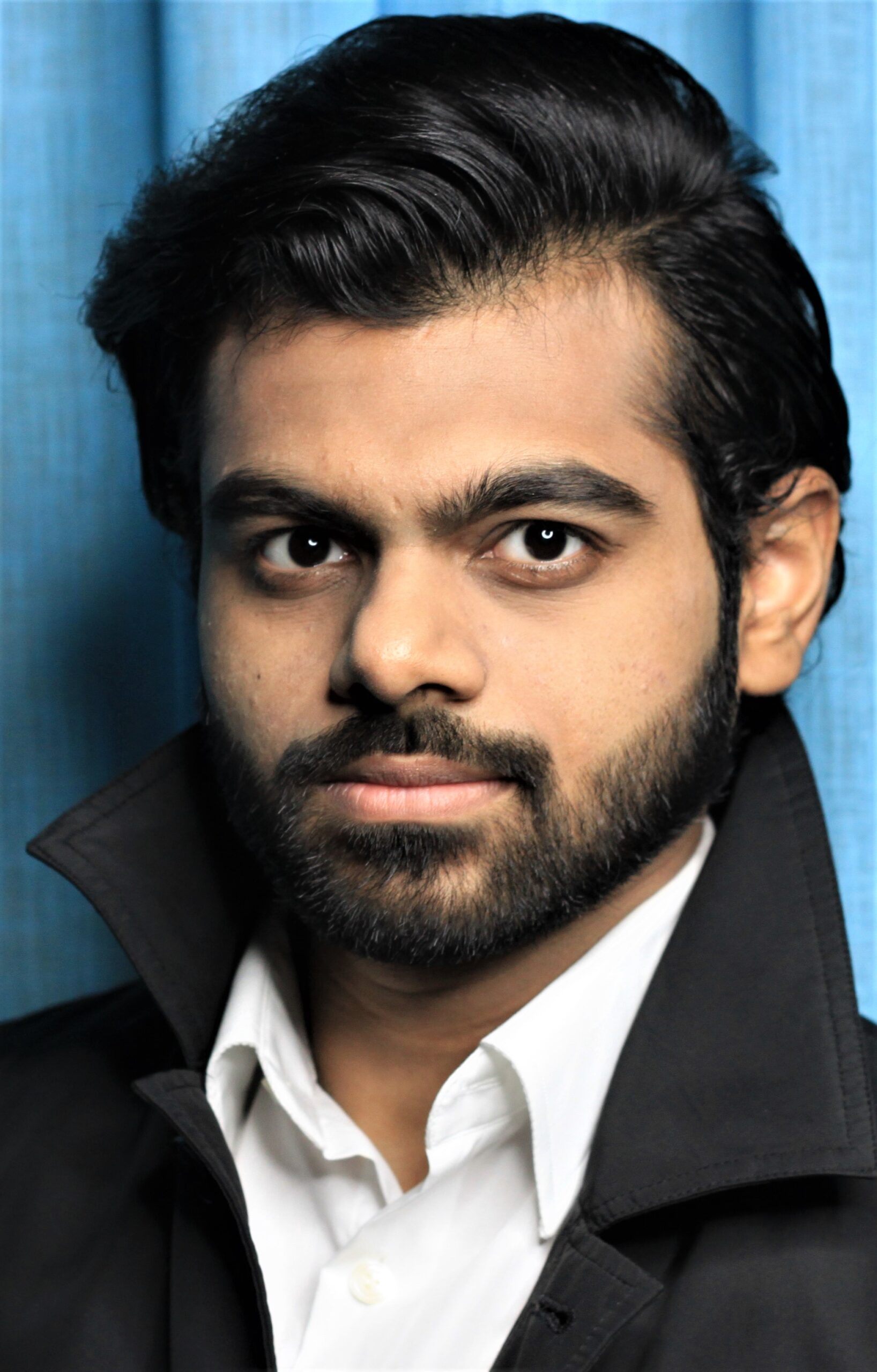 Me Standing in front of a blue background. I have jet black hair combed neatly to one side and a jet black beard of medium length (well groomed). I have olive skin, wearing a black trench coat over a white dress shirt.