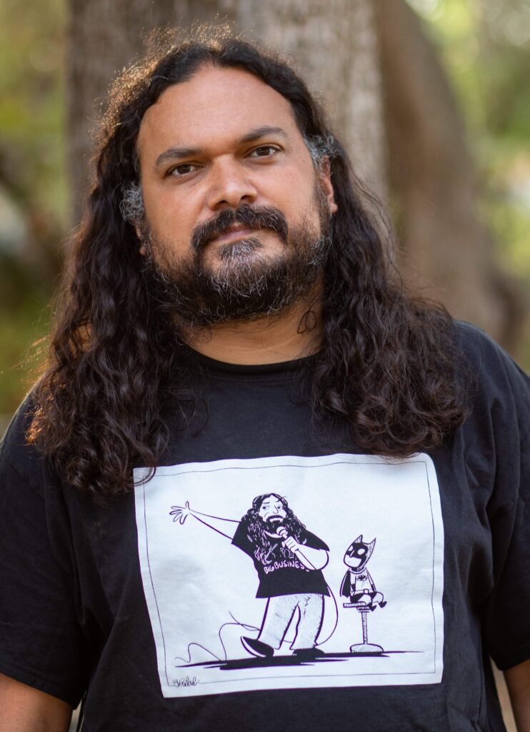 A Hispanic male with long black hair, a slightly graying black beard with a black shirt bearing a cartoon of himself telling standup comedy jokes stares intensely into the camera standing in a park