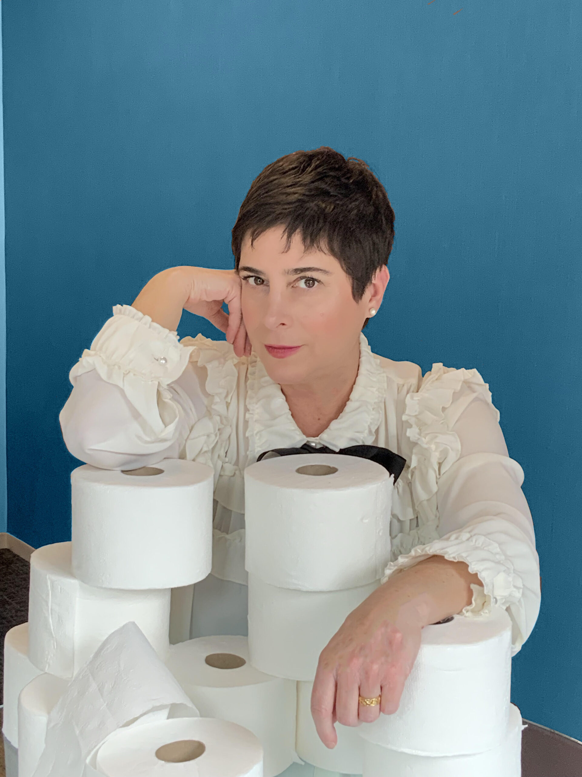 Amy is a smiling white woman with short black hair, wearing a white ruffled blouse. She is seated before a blue wall behind several stacks of toilet paper that she is leaning on.