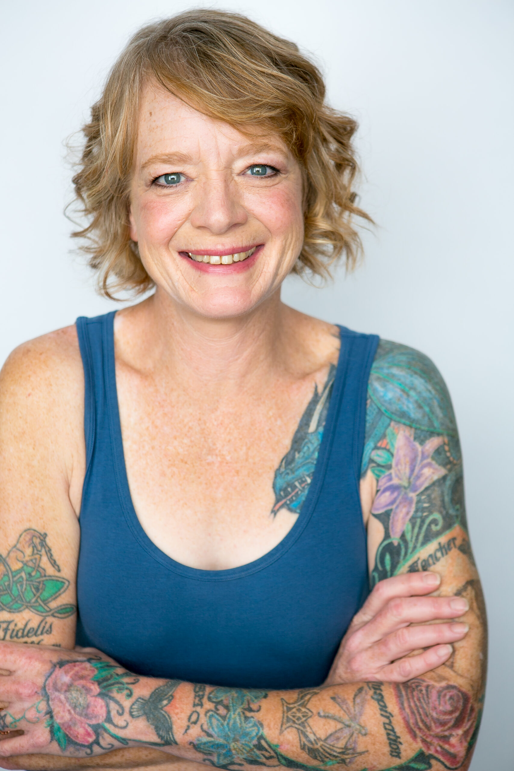 Gwyn is a middle aged woman with chin-length red hair and blue eyes. She is wearing a blue tank top that shows tattoos covering her left arm with smaller tattoos on her upper right arm and is standing with her arms crossed in front of her.