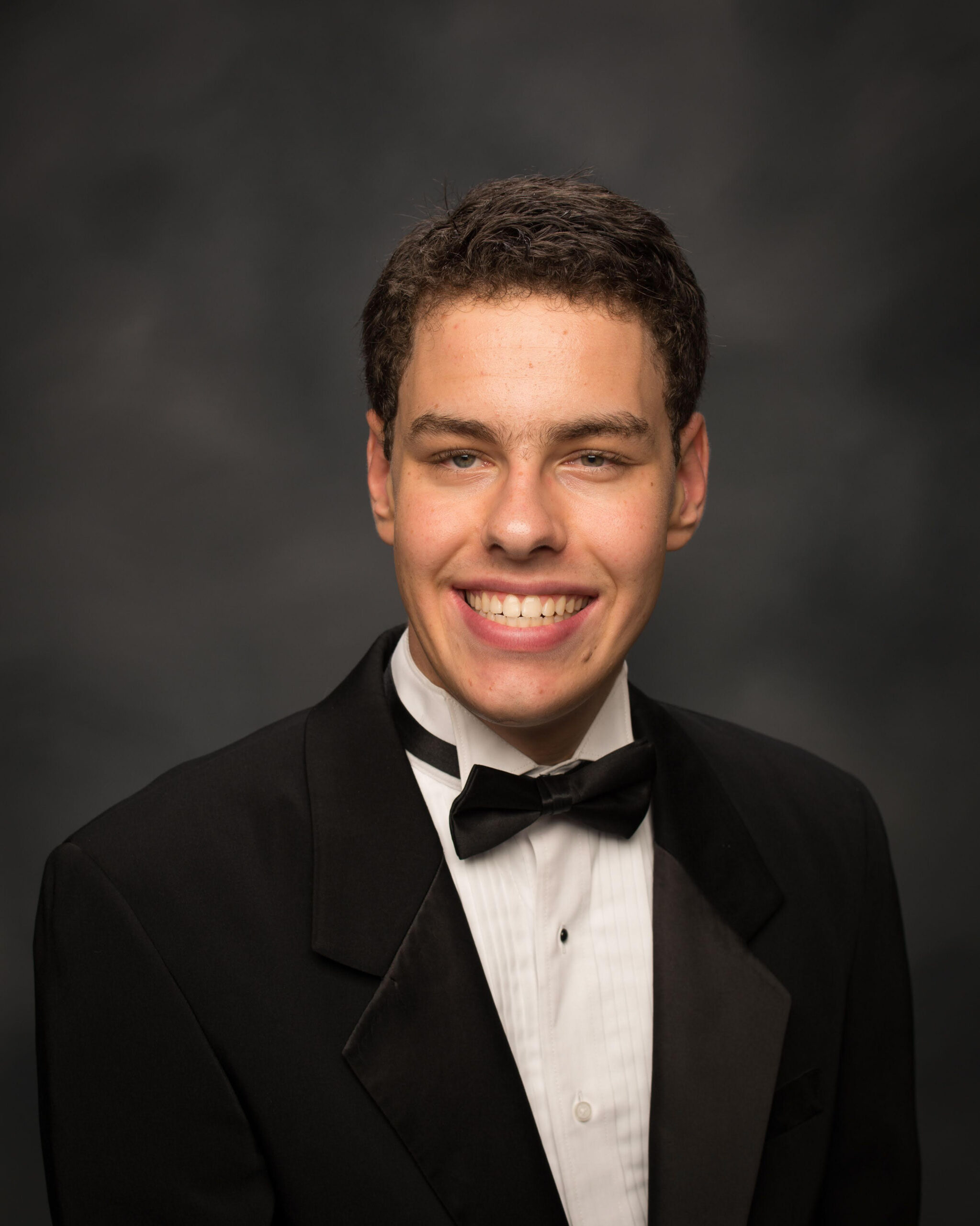 A brown-haired male in his early 20's wearing a black tuxedo jacket, white tuxedo shirt, and black bow tie. He is sitting in front of a dark gray background.