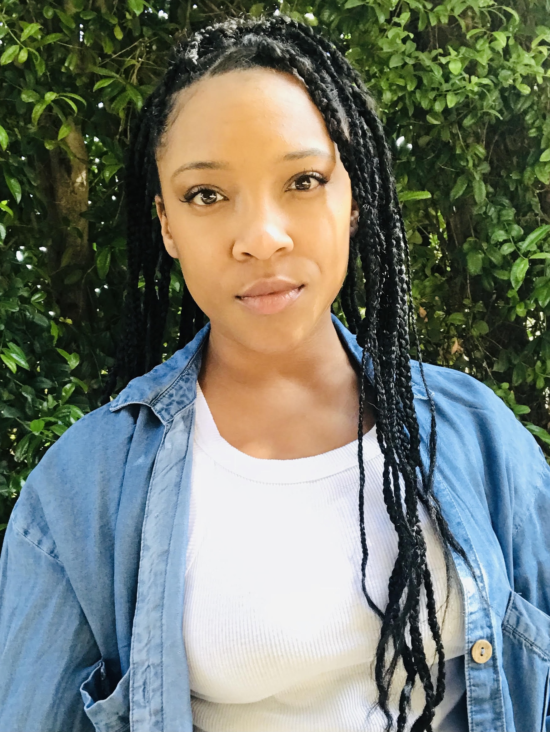 A Isrealite (Black) lady standing in her white tank top with her blue jean jacket on. She is smiling and glowing with her eyes staring straight to the camera and lots of crossroad triangle braids in individual hairstyles!