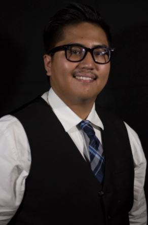 A brown, Filipino male with glasses wearing formal attire, which consists of a white shirt, blue tie, and black vest, in front of a black background..